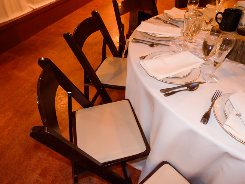 Several dark wooden garden folding chairs surrounding a round table set for a formal dinner covered with a white tablecloth and has plates, napkins, silverware, and glasses.