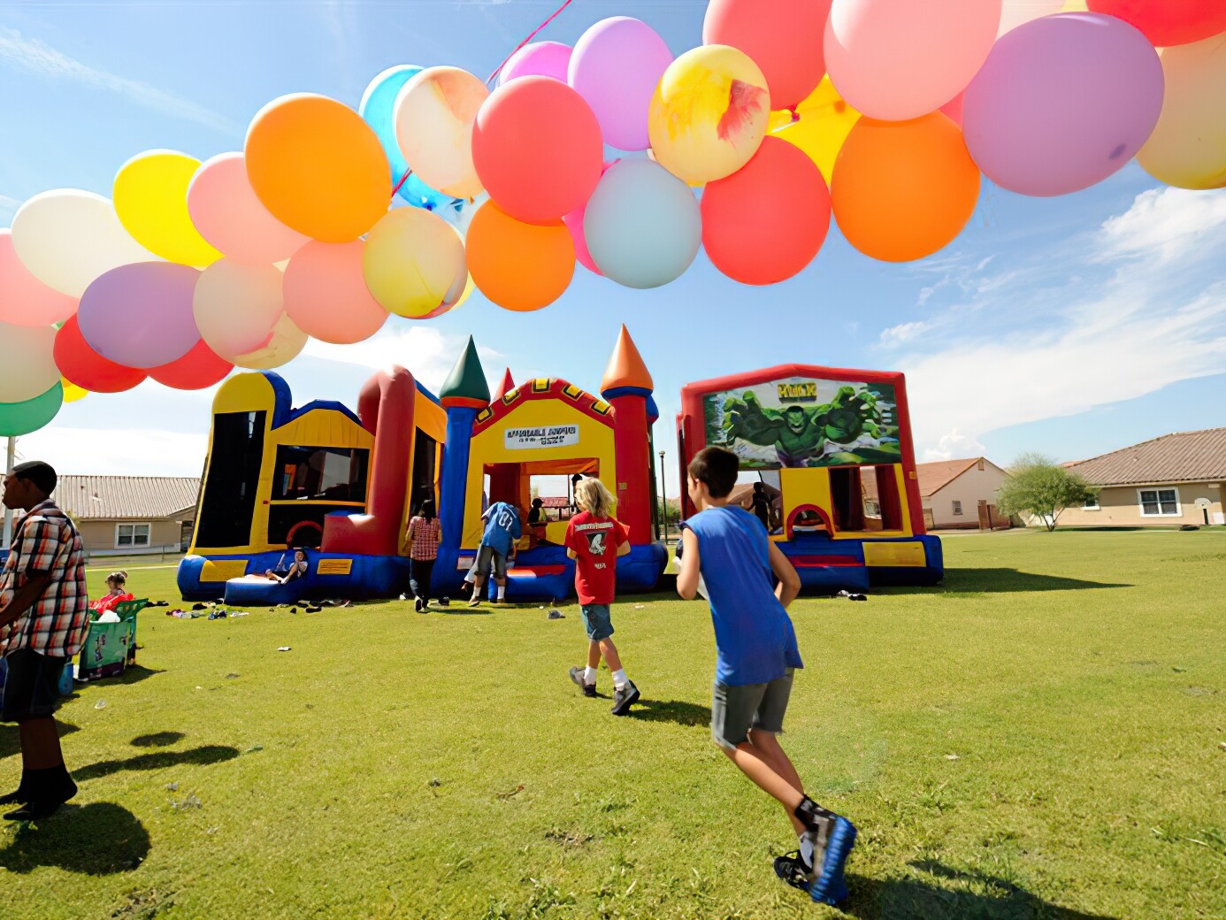Kids running under balloons to three inflatable bounce houses at a block party on a sunny day.