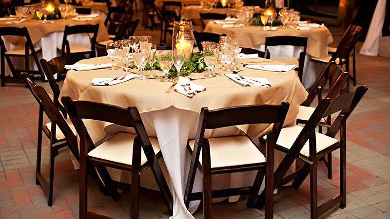 Large round table with eight brown and tan wooden chairs set with a white and tan table cloth for a nice wedding.