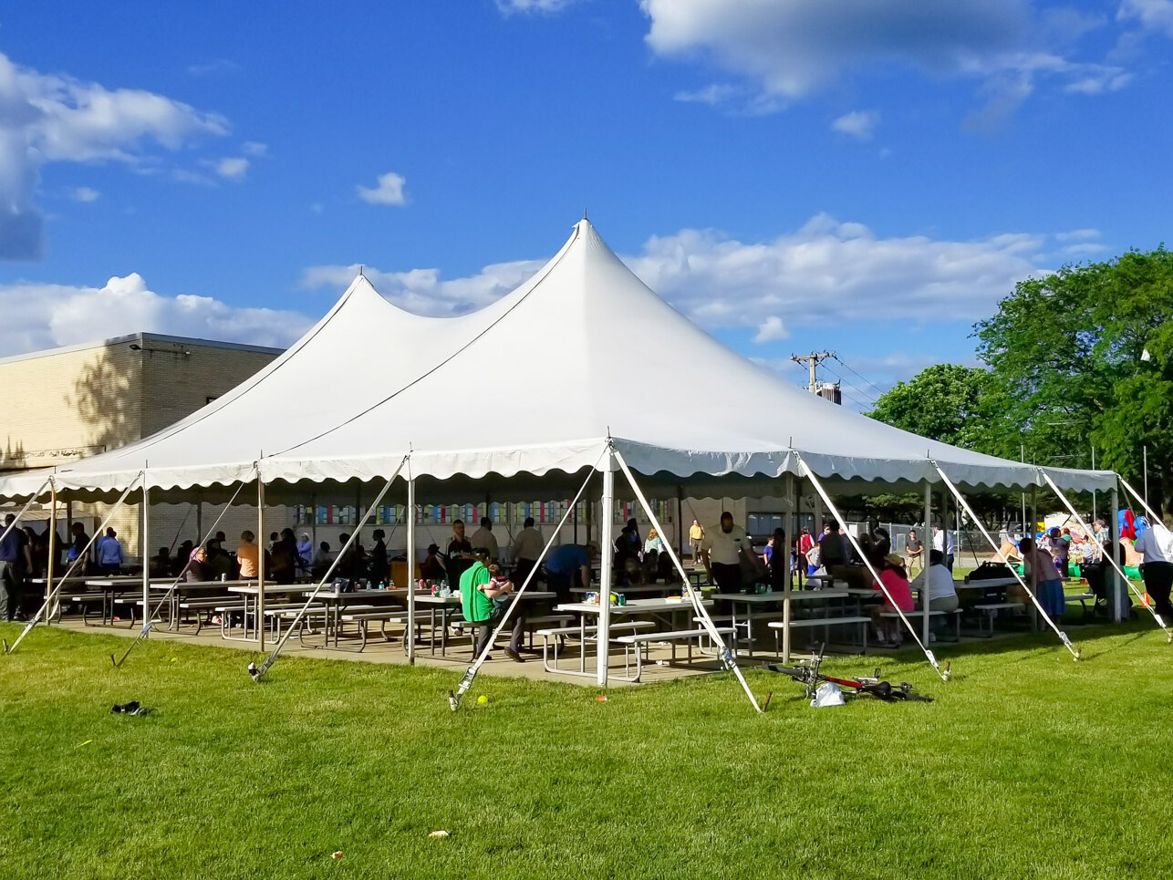 A high peak tent with picnic tables and people under it eating at a church picnic event.