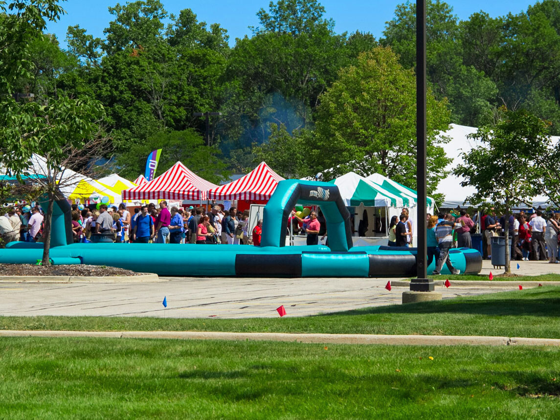 Multiple different color frame tents and an inflatable racetrack with many people outside at a corporate event party.