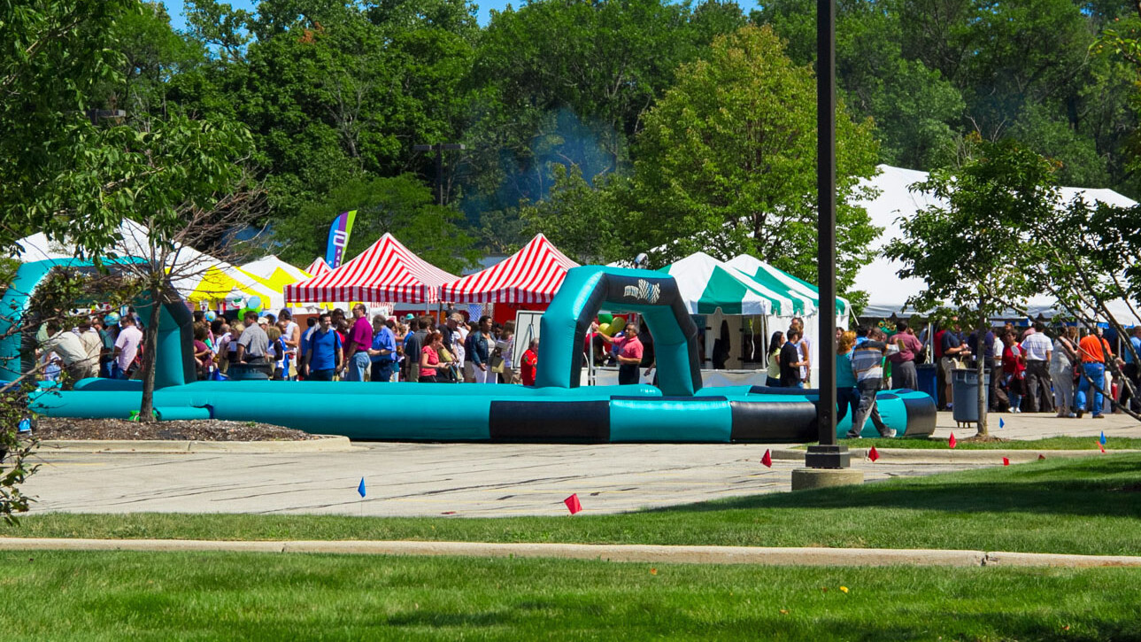 Multiple different color frame tents and an inflatable racetrack with many people outside at a corporate event party.