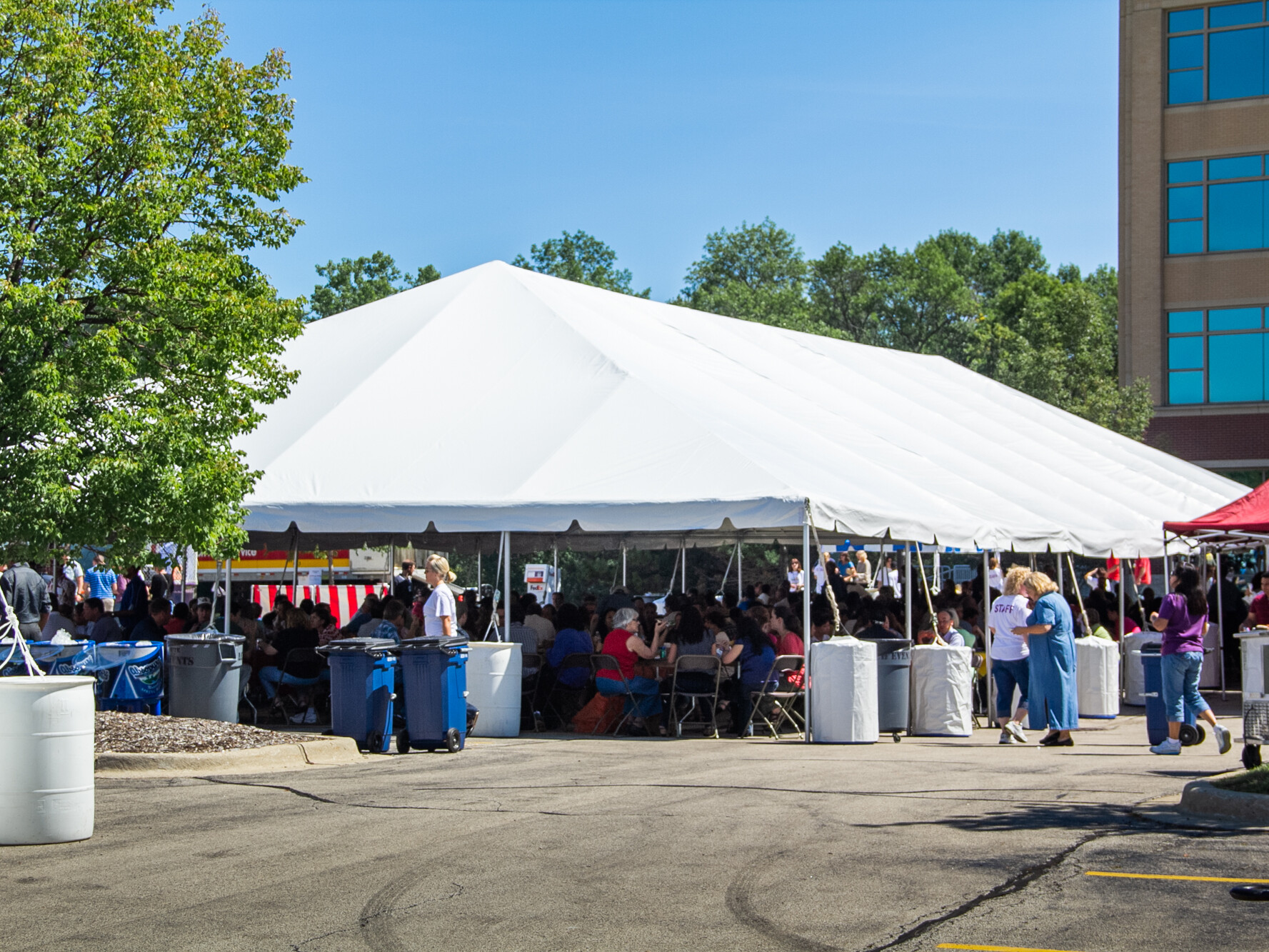 A massive frame tent in a parking lot with a lot of people sitting under it and eating at a corporate picnic.