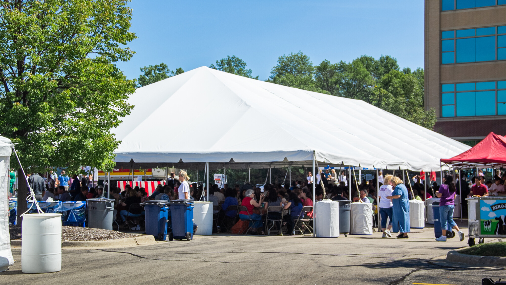 A massive frame tent in a parking lot with a lot of people sitting under it and eating at a corporate picnic.