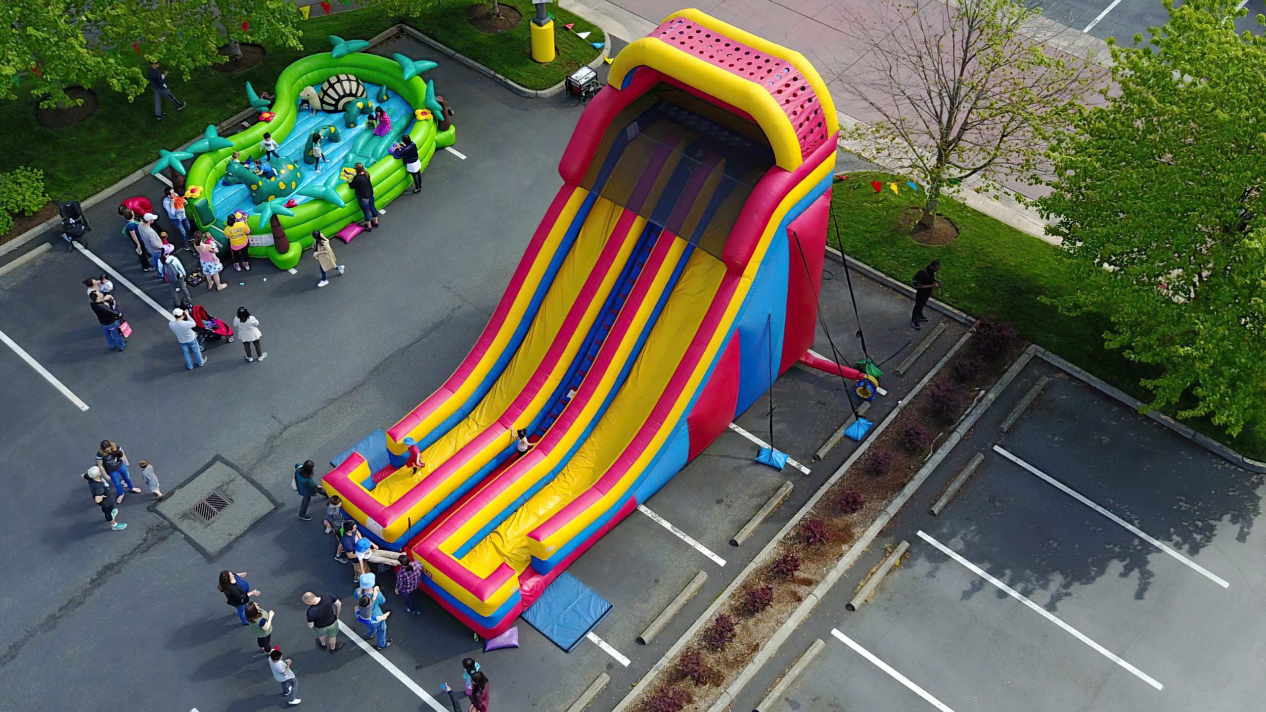 Overhead view of a giant slide in a parking lot next to a kids inflatable play jump and parents watching their kids play at an event.