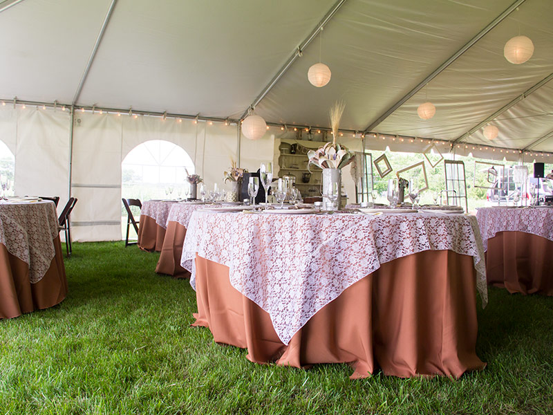 Tables under a frame tent with brown linen and a white flower pattern table cloth.