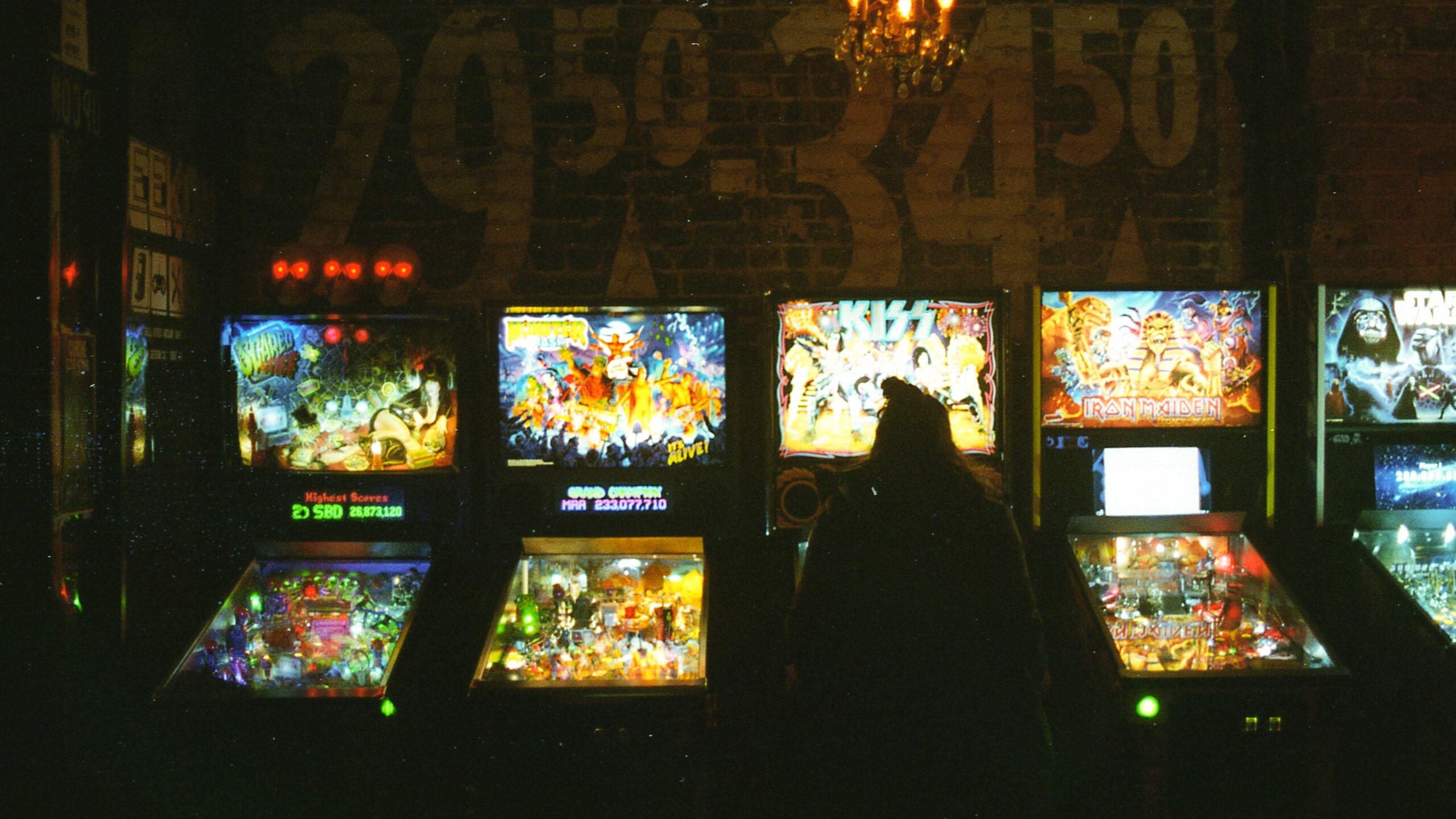 A row of lit up pinball games with a girl playing one in a dark room.