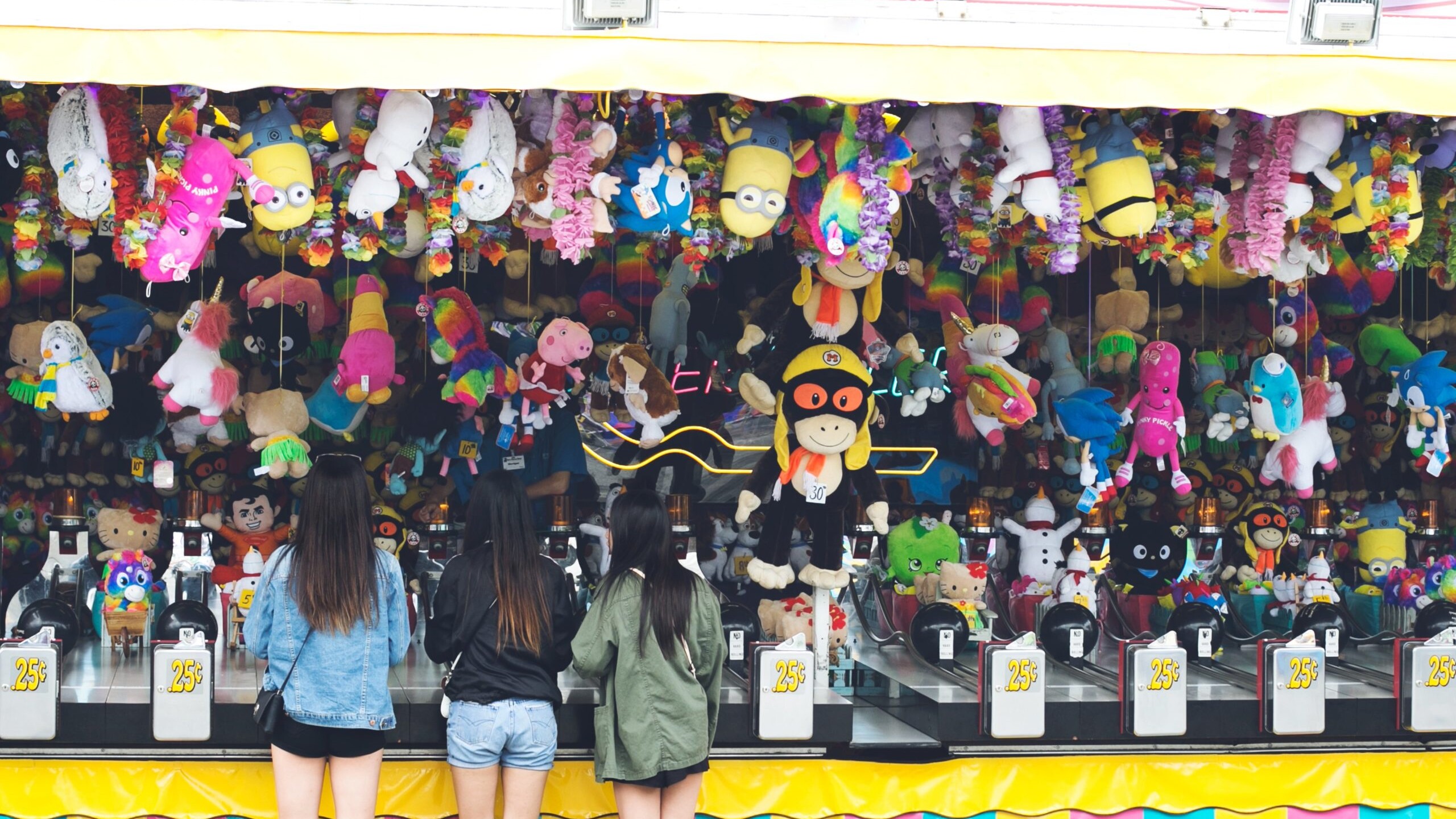 Three girls standing at a carnival game with a lot of toy prizes.
