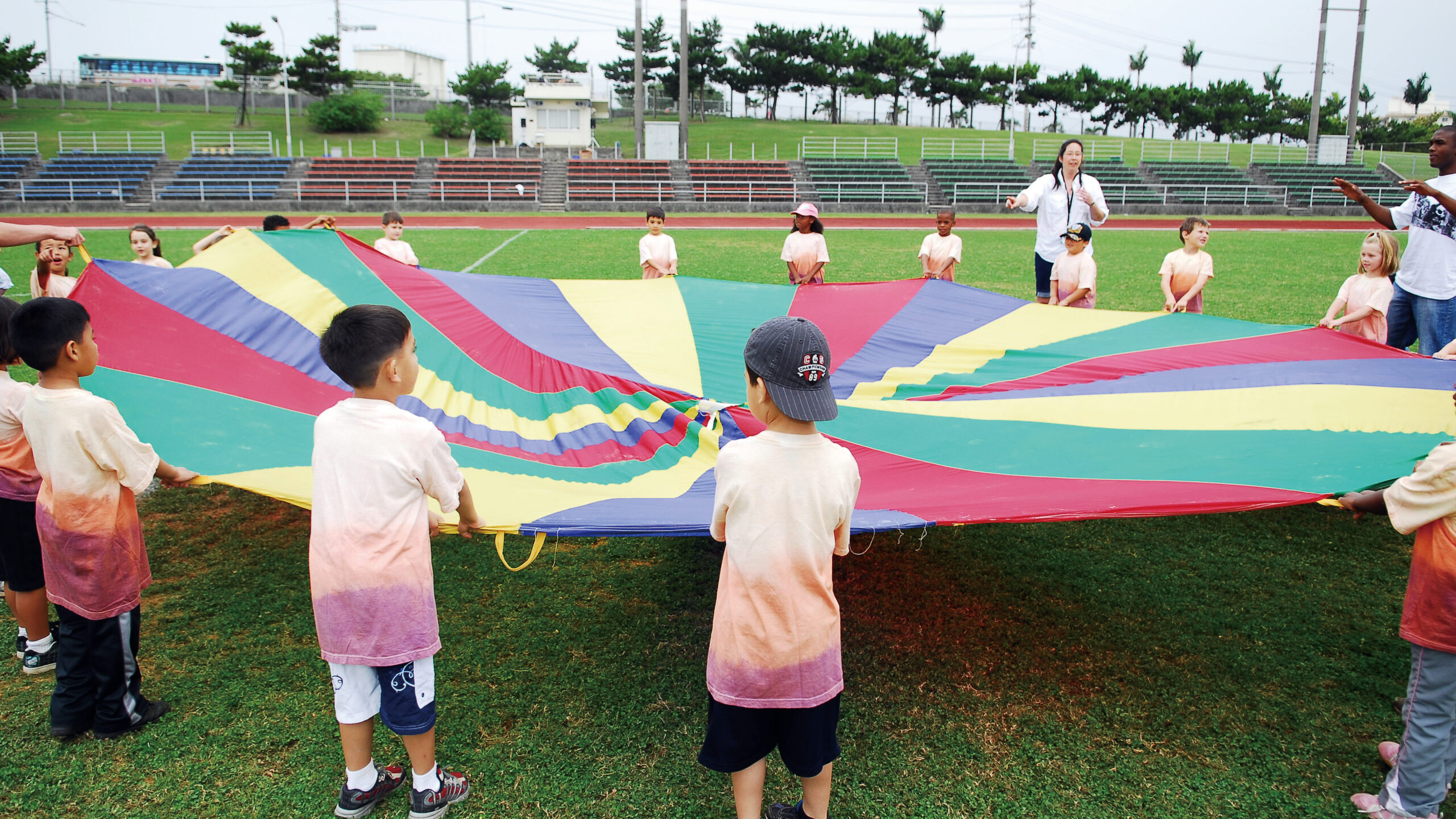Little kids holding a circular and colorful parachute with handles in a football field having fun for a school field day.