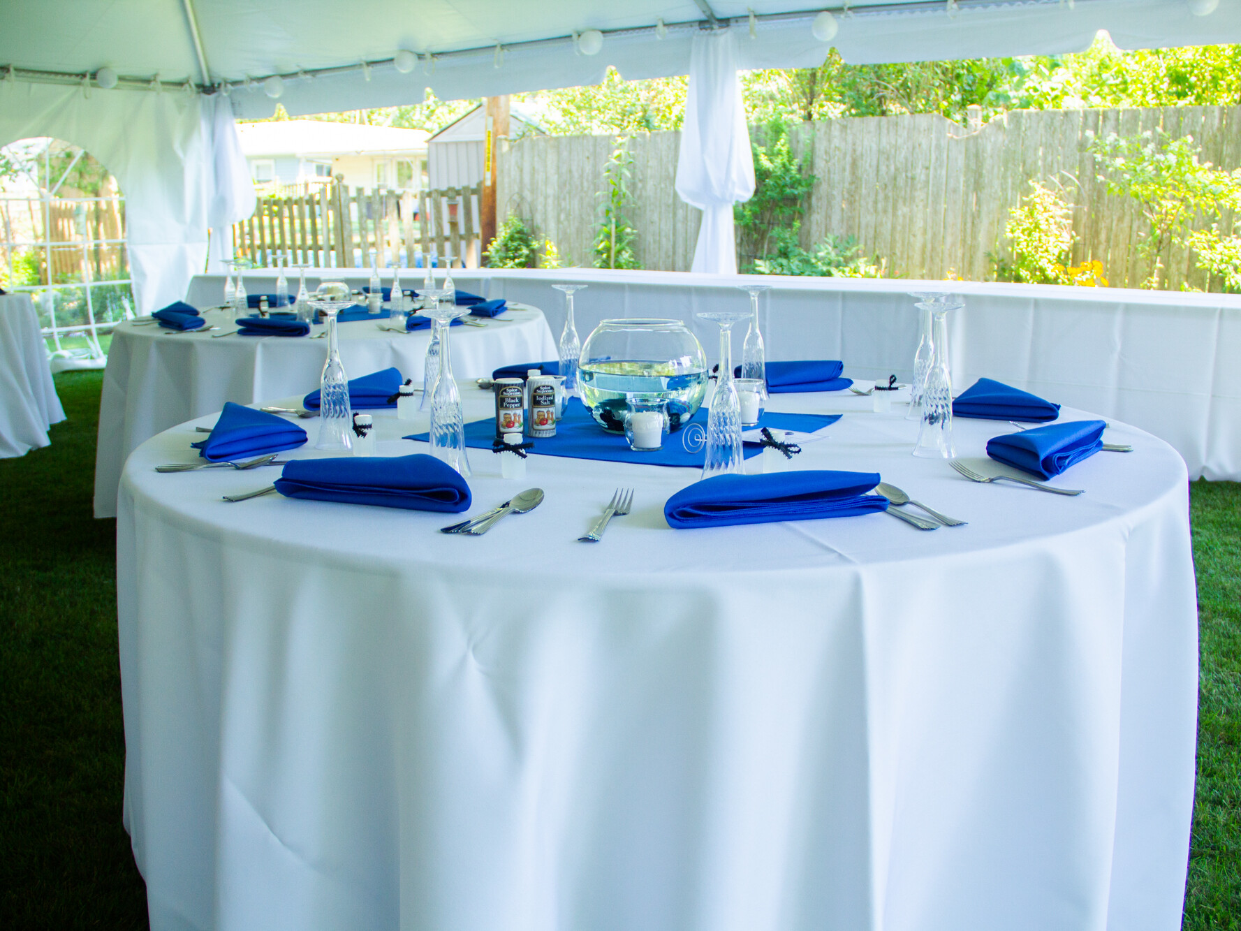Table set under a tent with a white tablecloth and blue napkins for a wedding.