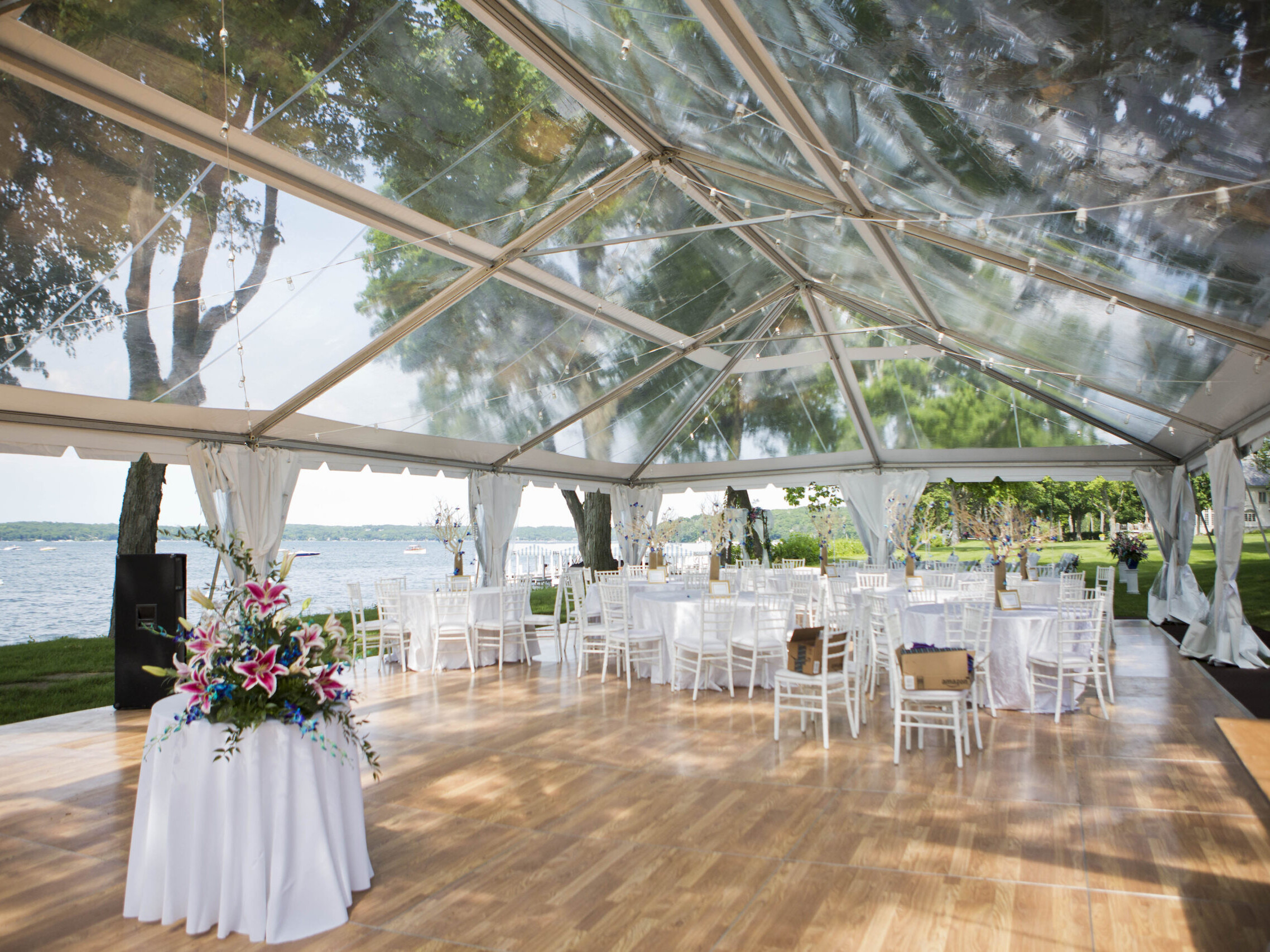 Inside a clear top tent on a hard wooden floor with tables and chairs set up outside on grass on a sunny day next to a lake for a wedding event.